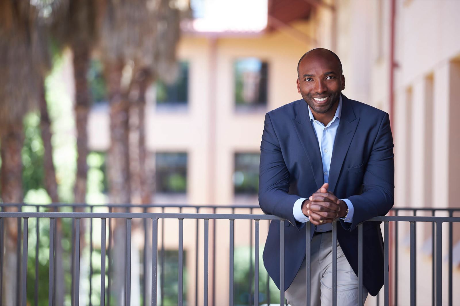 Man in suit leaning over railing and smiling at camera, palm trees in background. 