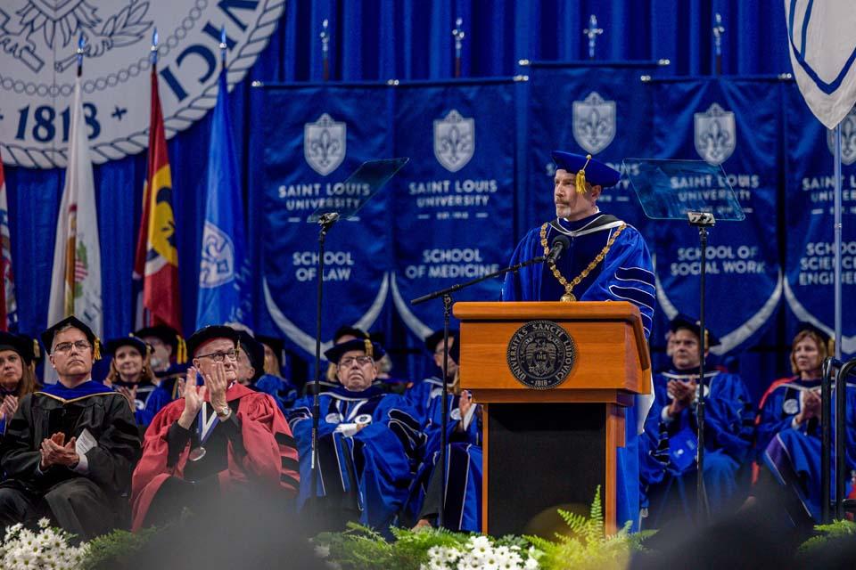 A man in a graduation cap and gown standing at a podium addresses an audience.