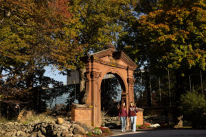 Two students in Ramapo College shirts walk toward the camera from the Havemeyer Arch on a fall day. The trees are covered in green, orange, and red leaves.