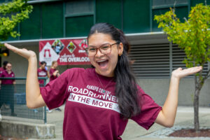 A student with long hair and glasses wears a maroon RCNJ t-shirt that says "Roadrunners on the move" and stands in front of The Overlook, a res hall on campus. She smiles and holds her arms out on each side of her in the air with enthusiasm.