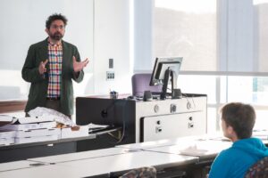 A man stands in front of a whiteboard talking with a computer to his right, while a student sits in front on him.
