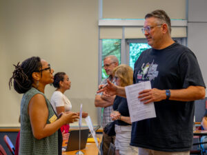 A woman wearing a green vest looks up at and speaks to a tall man wearing a blue T-shirt, holding a piece of paper.
