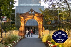Three students walked under the Havemeyer Arch with subtle sunlight rays cast between branches of trees. Maroon and white flowers line each side of Mansion Road in the foreground with tall grasses behind them.